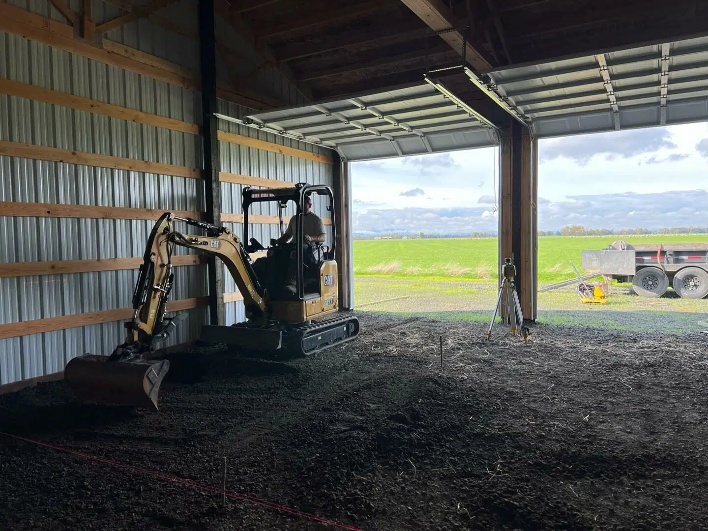 Excavator grading and leveling gravel inside a barn for shed pad preparation and sitework.
